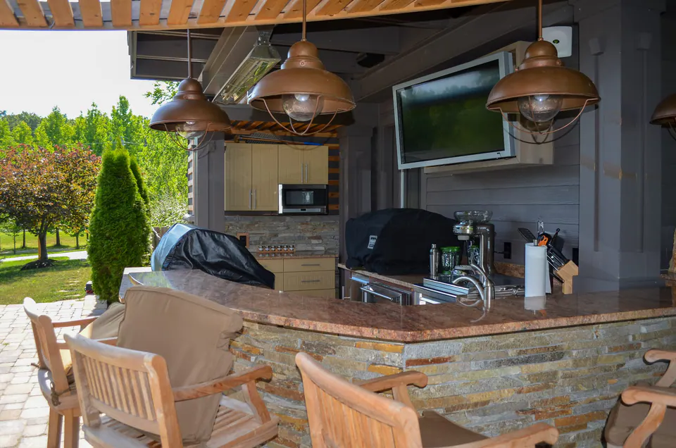 Outdoor kitchen bar under a covered roof with stone counter base, grill, sink, dining chairs, and pendant lights.